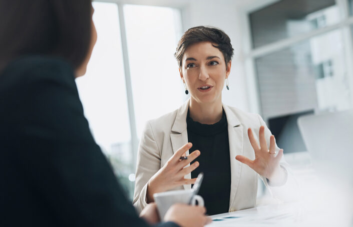 Stock image of a business woman wearing a black jumper and beige jacket having a one to one meeting with a colleague