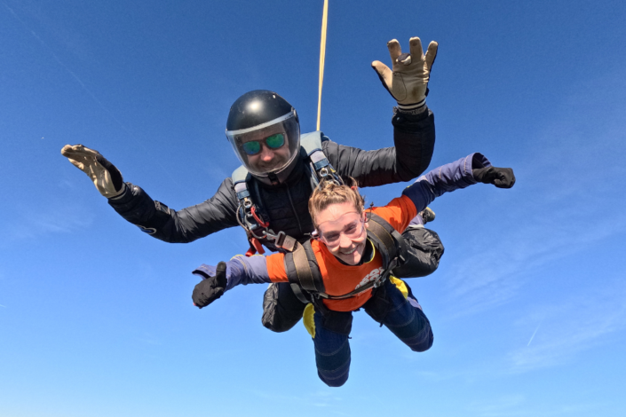 Photo of Char giving a thumbs up as she travels through the sky as part of a tandem skydive attached to the instructor with the blue sky in the background