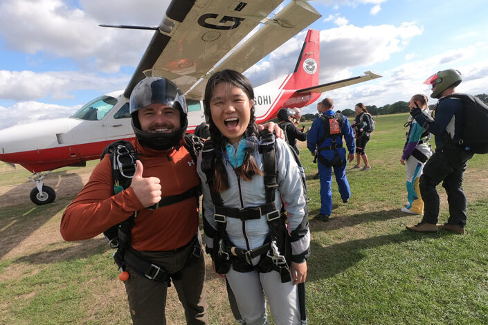 Photo of Jia standing next to her skydiving instructor with the plain behind her ready to start her skydive
