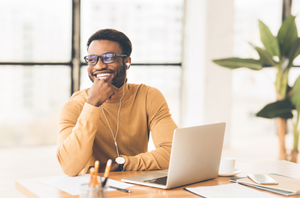 Photo of a man in his 30s wearing a yellow jumper sitting at a laptop looking happy